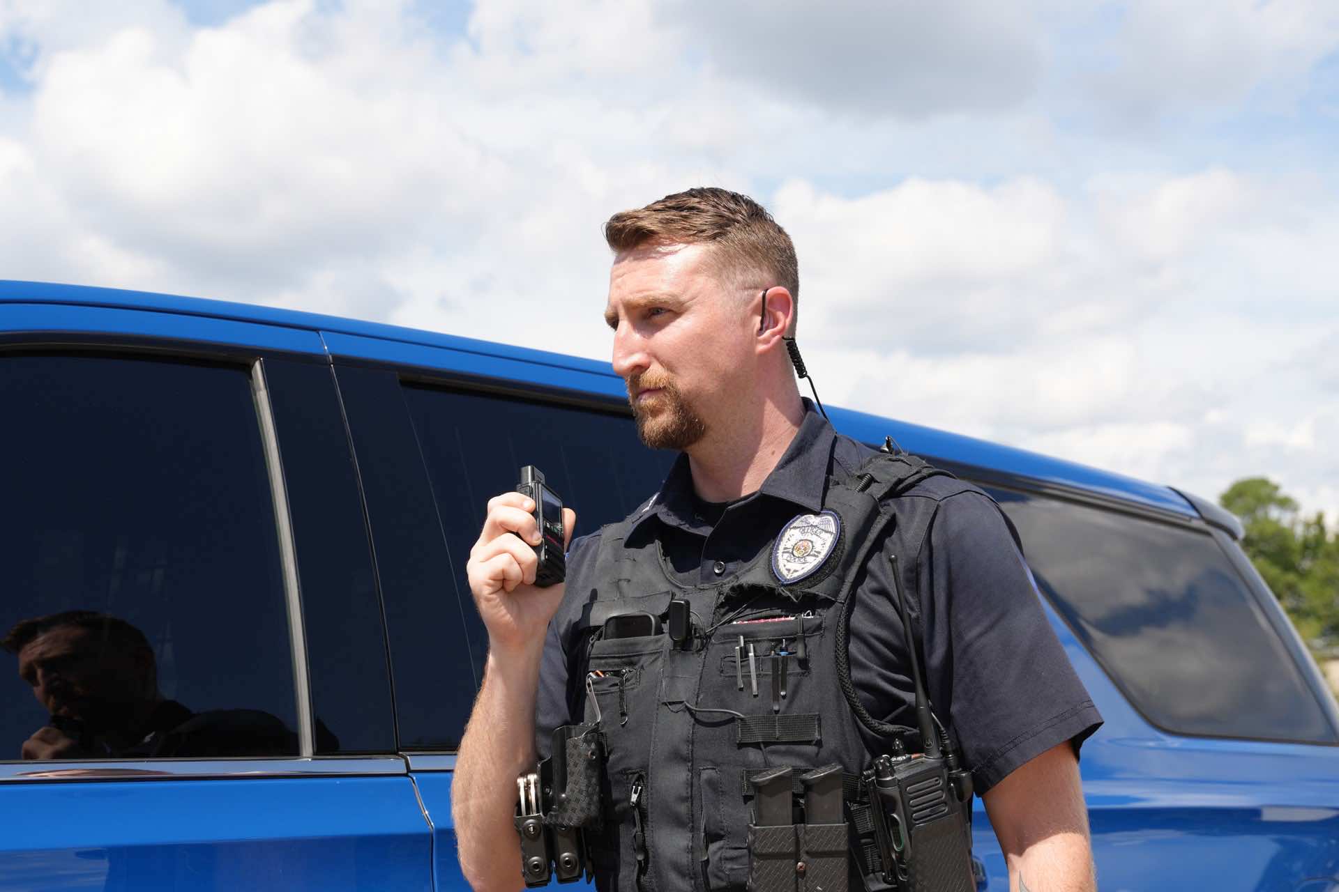 Police officer keying a SkyCom PTT radio beside a patrol vehicle