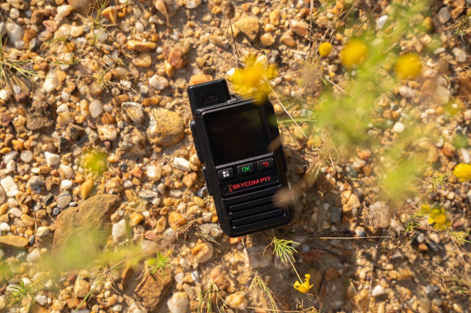 SkyCom PTT radio resting on gravel among wildflowers
